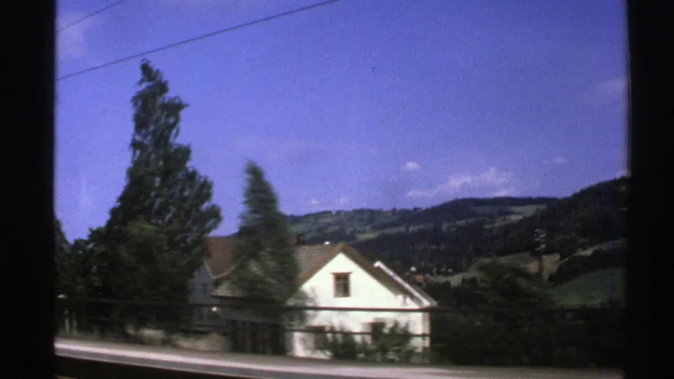 1976 Norway travel frame from a moving vehicle, with a white house, roadside trees, overhead wire, and green hillside landscape.