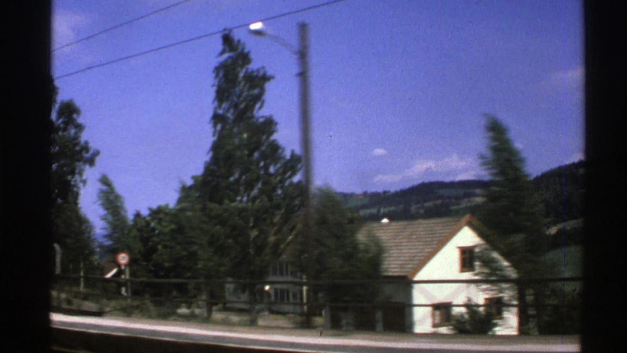 Roadside house, trees, fence, utility lines, and hills seen from a moving vehicle.