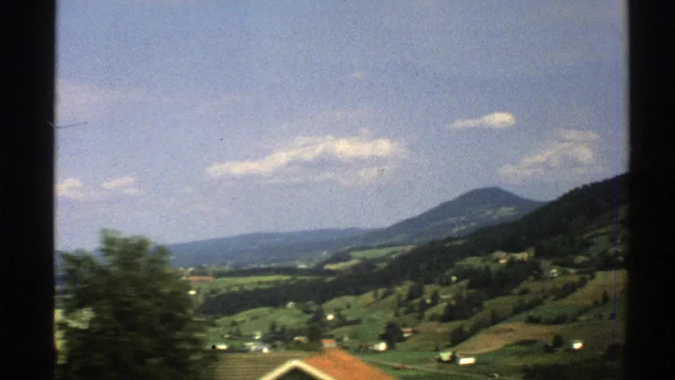 1976 Norway rural landscape with hillside fields, scattered farm buildings, trees, and a distant rounded peak.