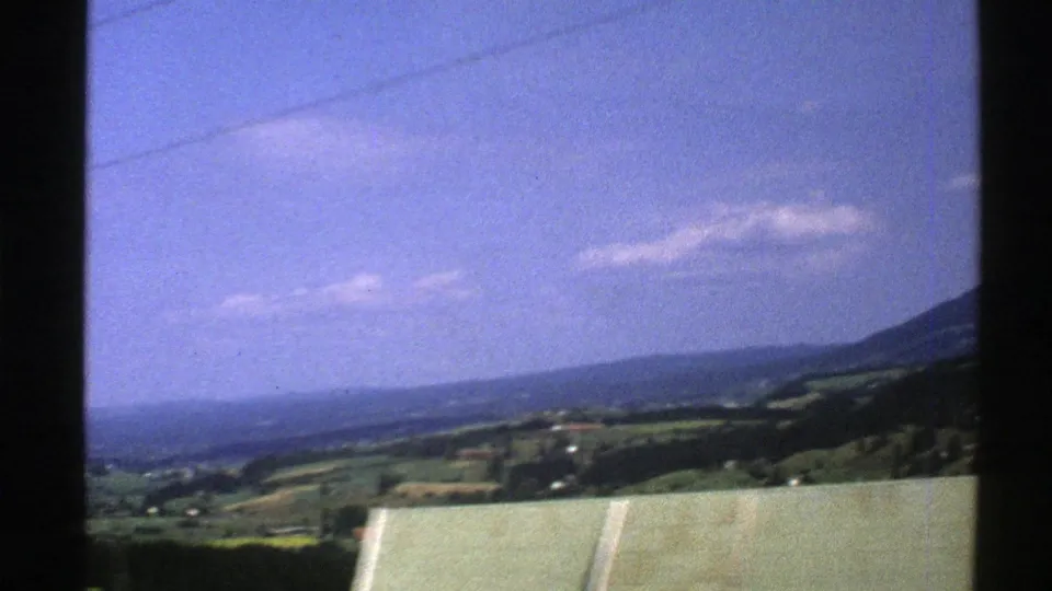 1976 Norway travel frame showing a broad valley, layered hills, fields, a roof edge, and utility lines under a bright sky.