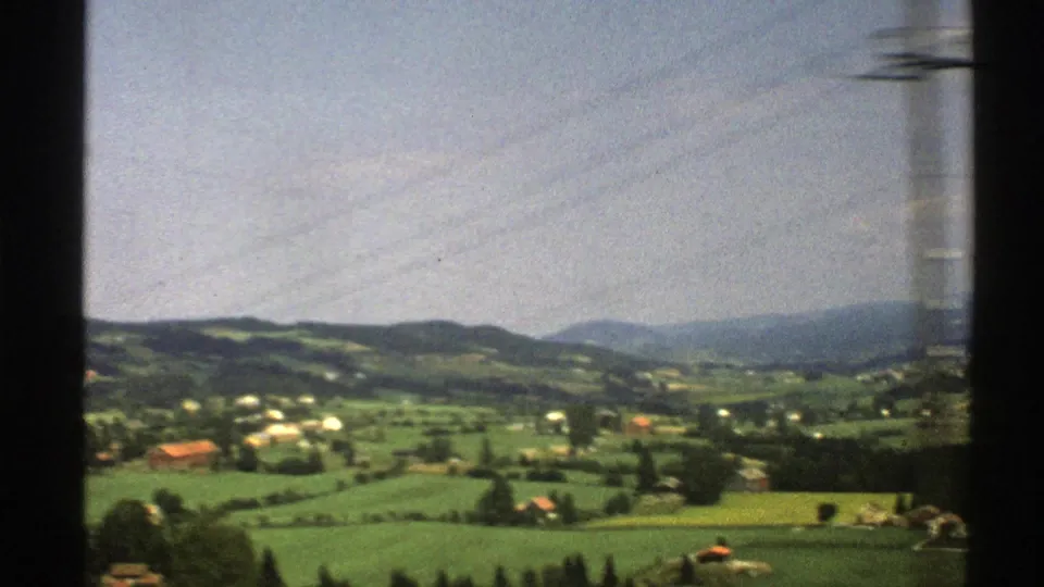 1976 Norway rural landscape with fields, small buildings, wooded slopes, and a blurred roadside pole from the moving vehicle.