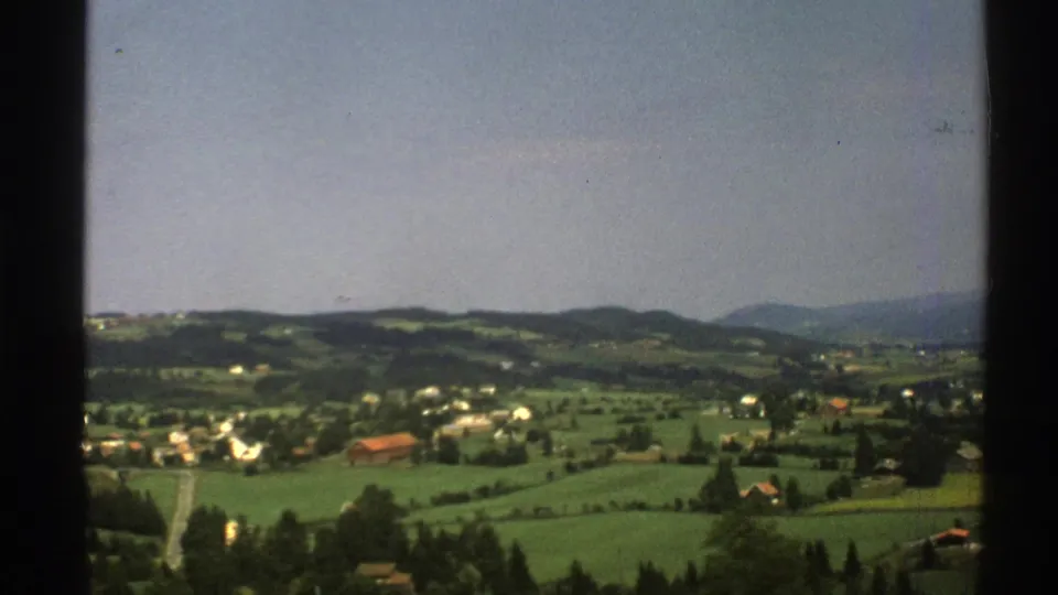 A 1976 Norway home-movie frame shows a rural valley with fields, scattered buildings, a road, and distant hills.