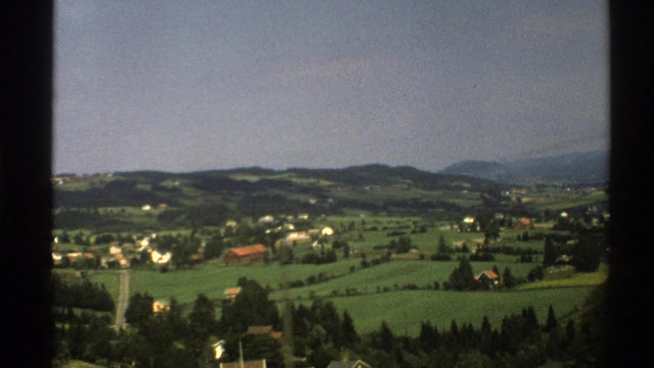 Green Norwegian countryside with scattered houses, fields, hills, and window-frame edges.