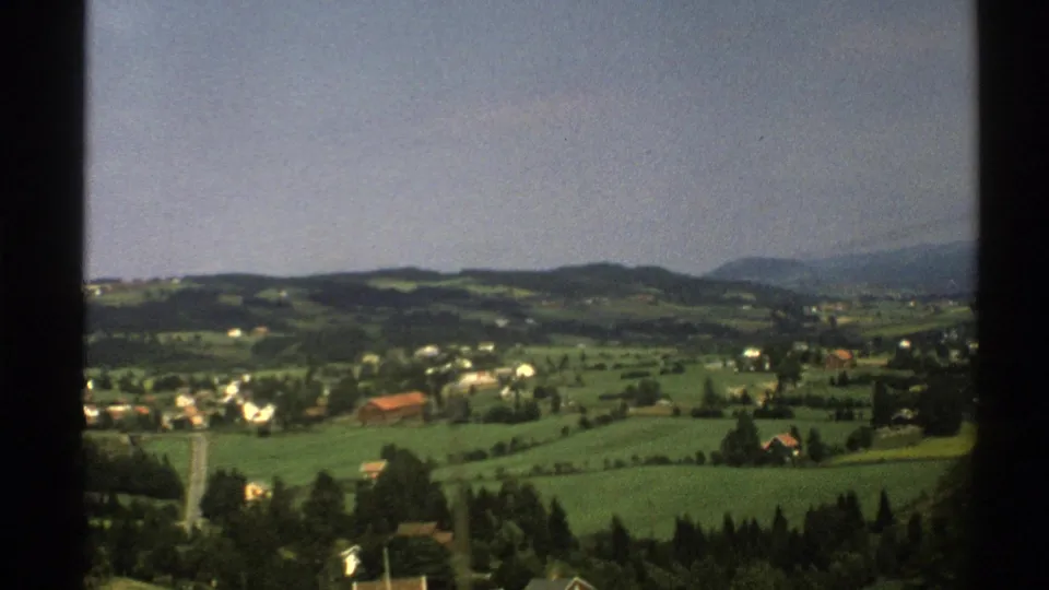 1976 8mm view across Norwegian countryside with green fields, scattered houses, red-roofed buildings, and distant hills.
