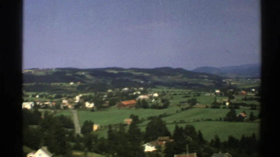 1976 8mm frame of Norwegian rural terrain with a road, fields, houses, tree cover, and hazy hills beyond.