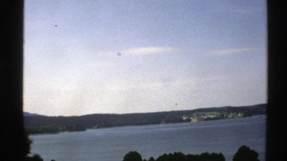 1976 Norway home-movie view of a broad lake or fjord with a wooded shoreline, low hills, and pale blue sky.