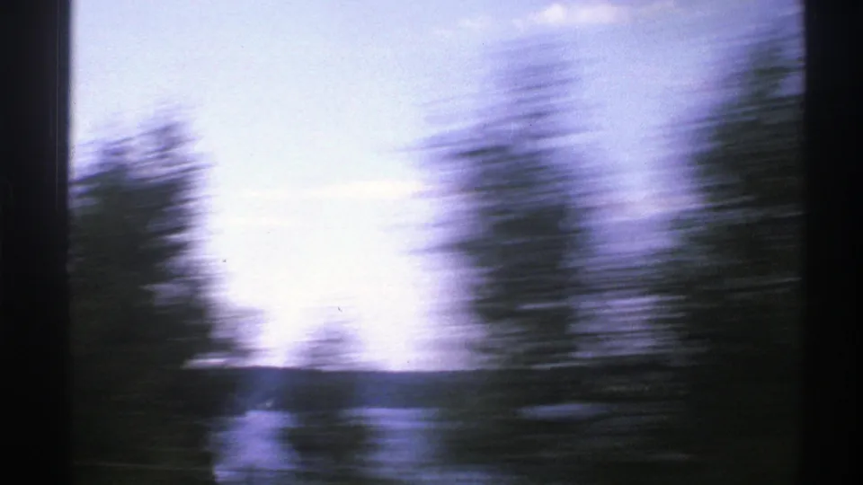 Motion-blurred trees sweep across a 1976 Norway home-movie view of blue water, low hills, and bright daylight.