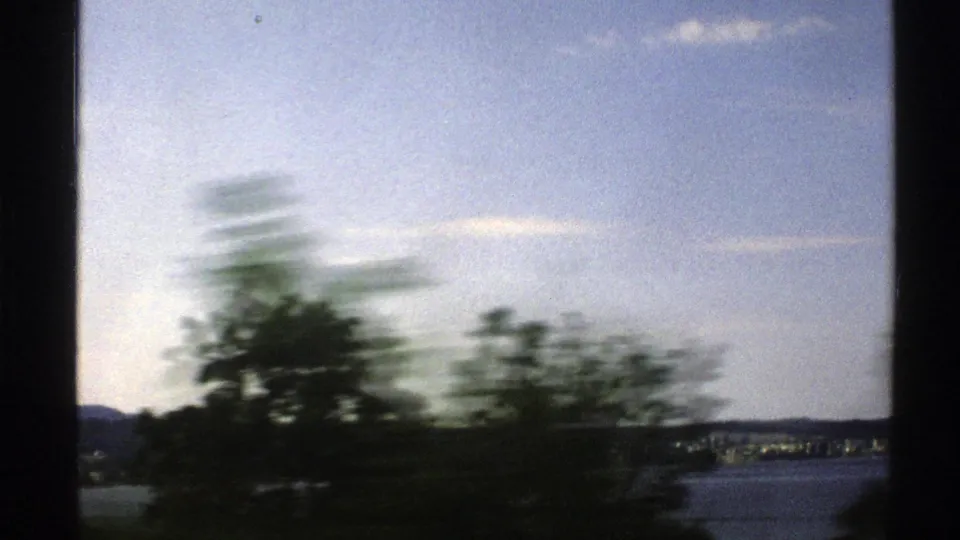 Moving 8mm view in Norway with foreground trees, blue water, a distant shoreline, low mountains, and pale clouds at the end of the clip.