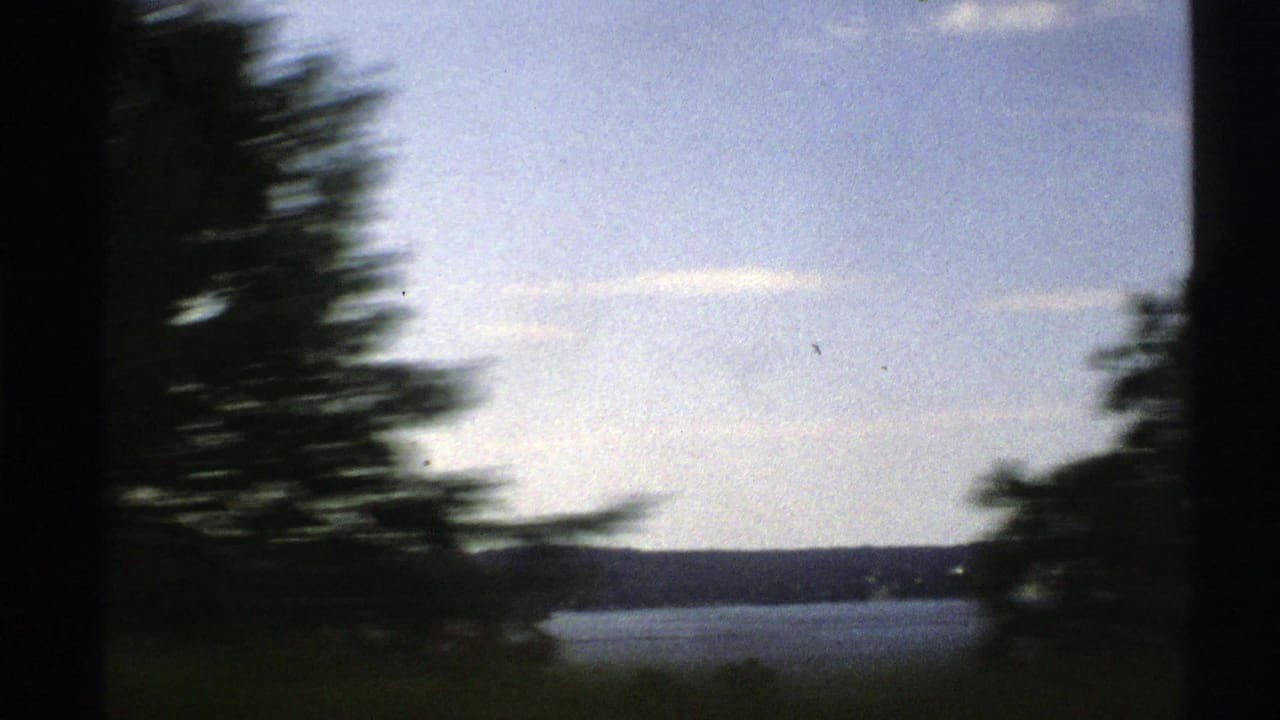 1976 Norway home-movie view from inside a moving car, with dark window framing, blurred trees, water, and low hills beyond.