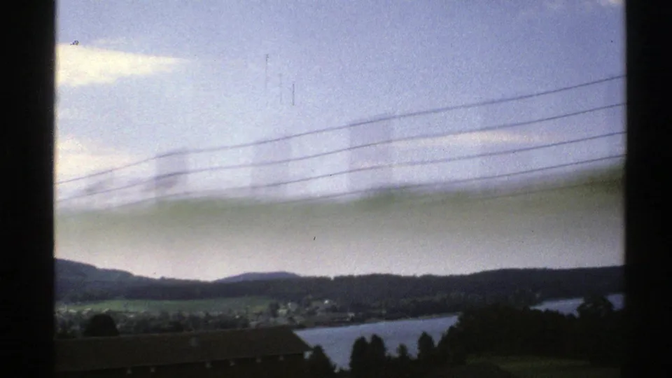 Passenger-window view of rural Norway in 1976, with a lake, low hills, rooftops, power lines, and blurred grass crossing the foreground.