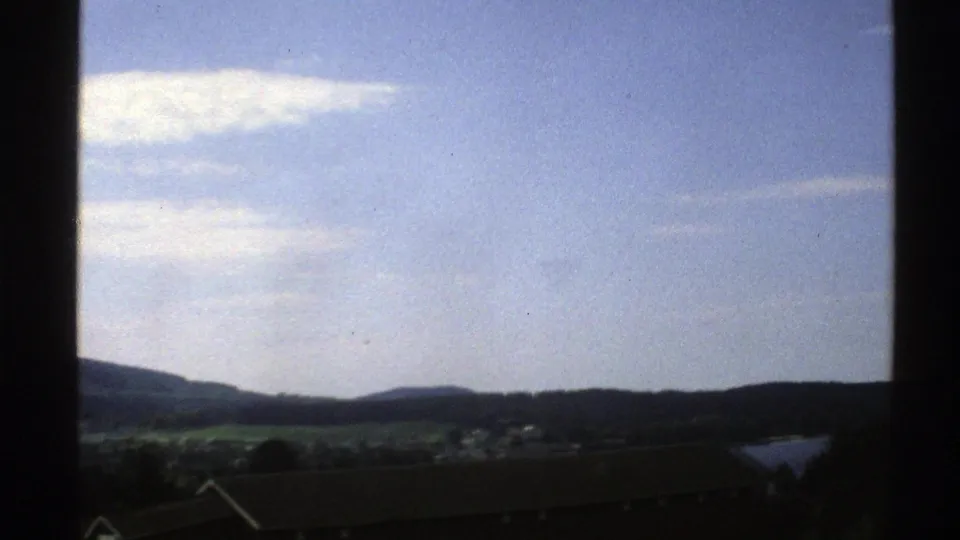 1976 Norway travel footage frames an open lakeside landscape with fields, low hills, rooftops, clouds, and dark vehicle-window borders.