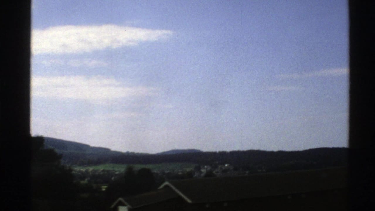 1976 Norway 8mm view of blue sky, low clouds, distant hills, and rooftops seen through a dark window frame.