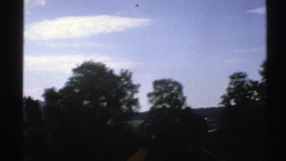 1976 8mm frame showing shadowed trees against a pale blue sky, with hills and small distant structures beyond.