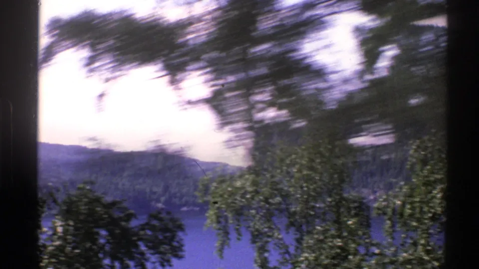1976 Norway home-movie frame with roadside trees sweeping across a partial view of a lake and forested hillside.