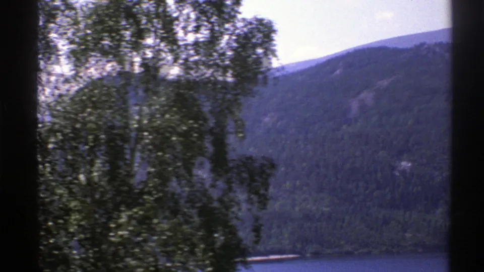 1976 Norway landscape frame with foreground trees, a forested hillside, and a narrow lake edge visible below.
