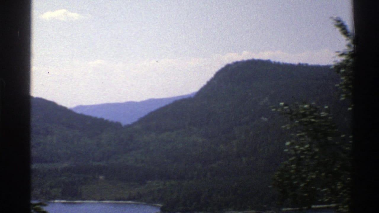 A 1976 Norway travel view across a waterway toward layered forested hills, with nearby trees blurred by motion.