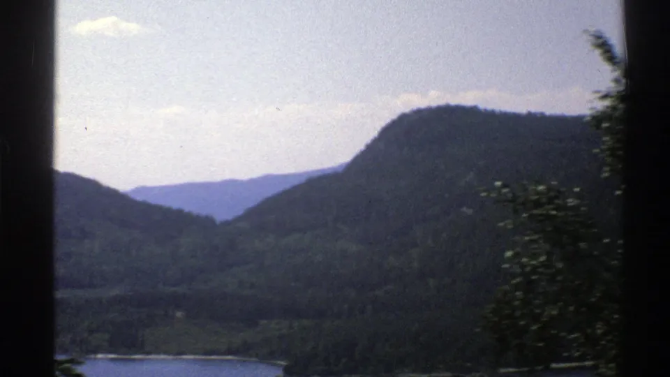 A 1976 Norway travel view across a waterway toward layered forested hills, with nearby trees blurred by motion.