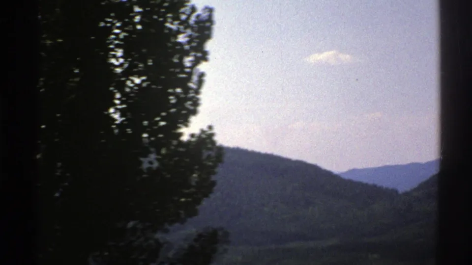 1976 Norway 8mm travel frame showing layered forested hills under a pale sky, partly obscured by a dark roadside tree and vertical edge.