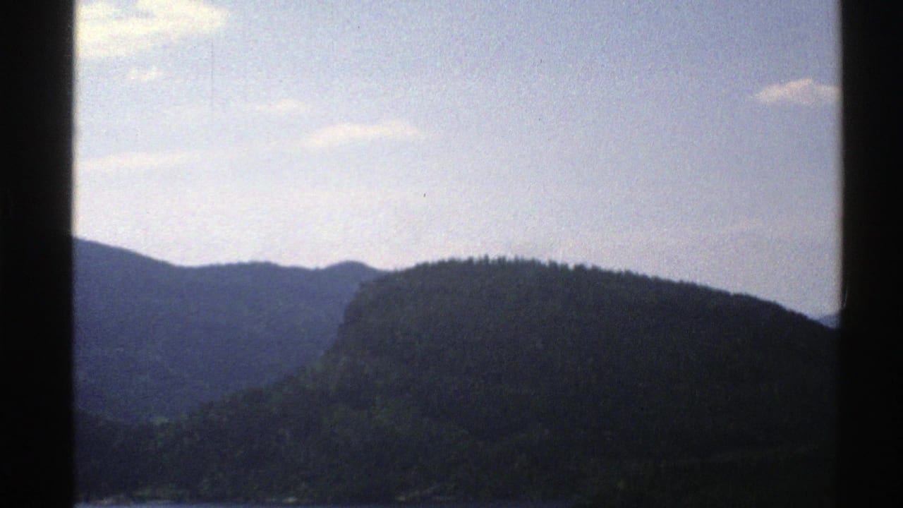 1976 Norway 8mm frame with blue-green ridges and forested slopes under scattered clouds, split by a dark vehicle or window edge.