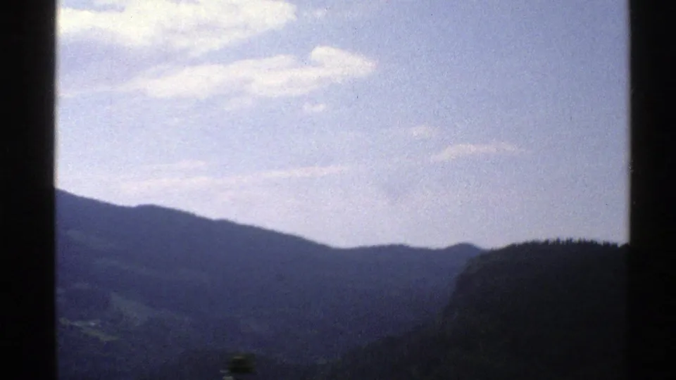 1976 Norway 8mm frame showing rolling mountain ridges and a dark forested hill beneath a pale blue sky with scattered clouds.