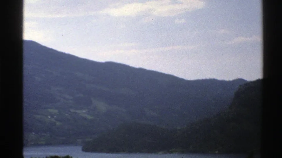 Norwegian mountain landscape and waterway seen from a moving vehicle or train, with a dark vertical window post in view.