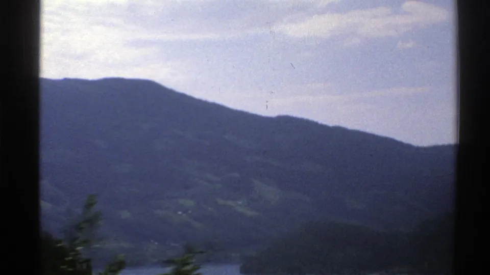 1976 Norway home-movie frame showing distant mountains, patches of green hillside, water below, and dark window framing.