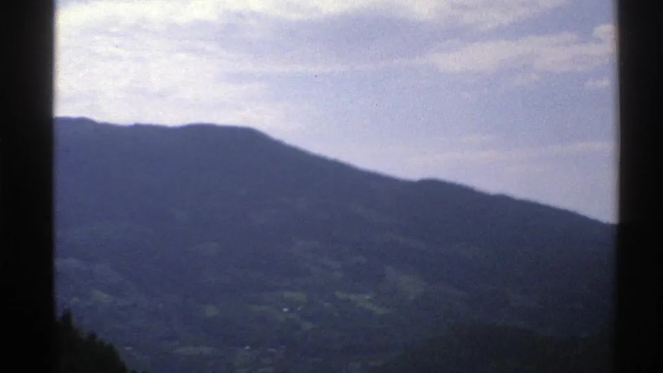Soft 8mm view of Norway's mountainous countryside with a broad ridge, green valley patches, and pale cloud cover.