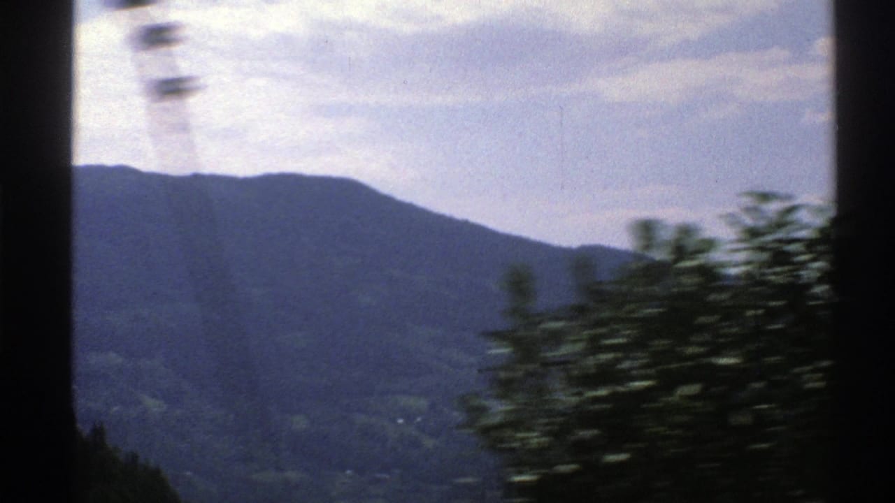 View through a vehicle window toward forested mountains and a cloudy sky.