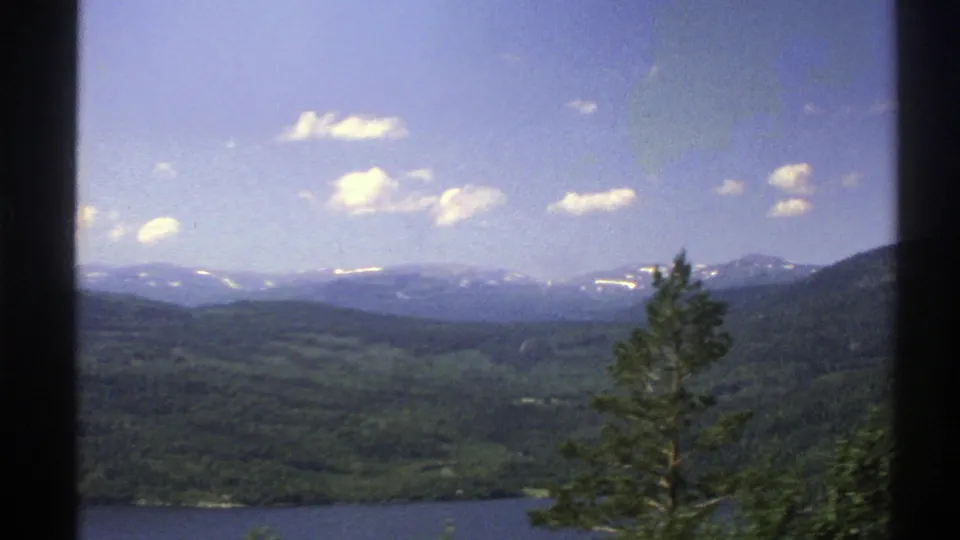 1976 Norway 8mm frame showing a broad green valley, a dark waterline, distant snow-streaked mountains and scattered clouds.