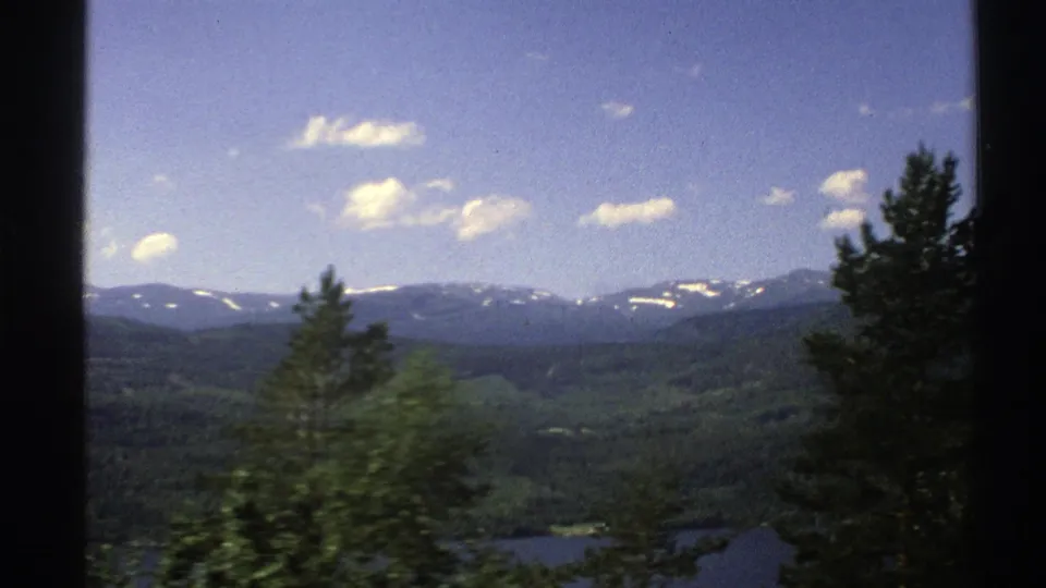 A moving 1976 Norway landscape view shows window framing, foreground trees, a strip of water and distant snow-dusted mountains.