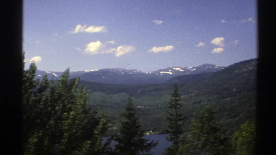 1976 Norway 8mm frame showing a broad green valley, a lake edge, dark conifers, and snow-streaked mountains beneath scattered clouds.