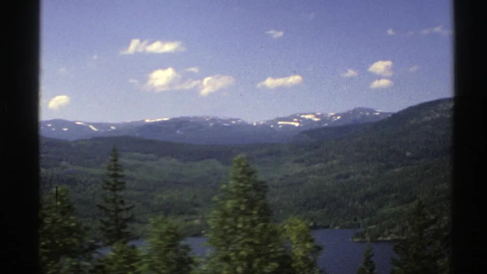 1976 Norway 8mm frame ending on a lake bordered by forested hills, foreground trees, and a distant snow-dotted mountain range.