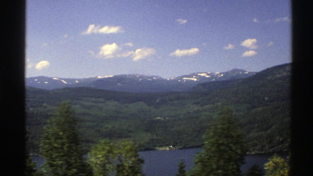 Blue lake below forested hills with snow-dusted mountains in the distance.