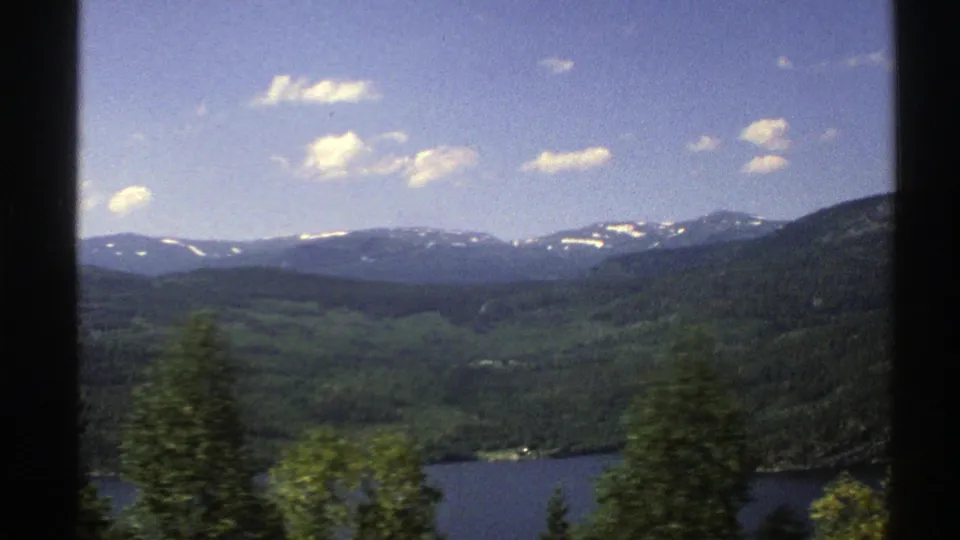 1976 Norway 8mm frame showing a blue lake, green forested valley, and distant snow-dusted mountains under a clear sky.