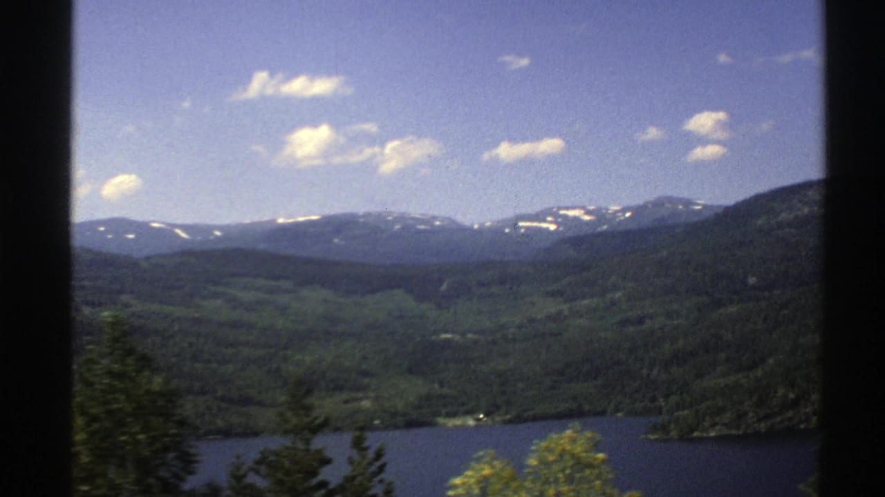 Forested valley and lake with distant mountain ridges under a blue sky.