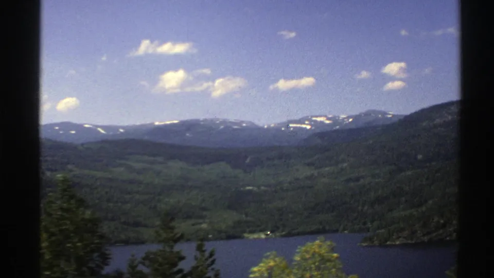1976 Norway landscape frame with dark tree slopes, a lake edge, and hazy snow-marked mountains beyond the valley.