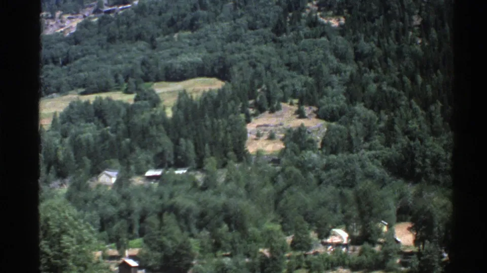 1976 Norway 8mm view looking down over dense trees, pale fields, and scattered rural cabins on a hillside.
