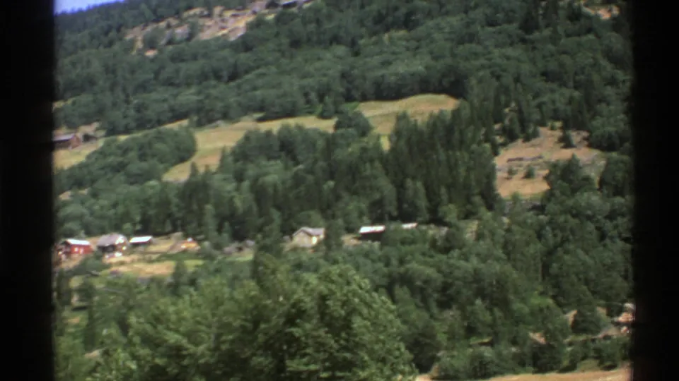 1976 Norway home-movie frame showing a rural valley hillside with cabins, fields, and surrounding forest.