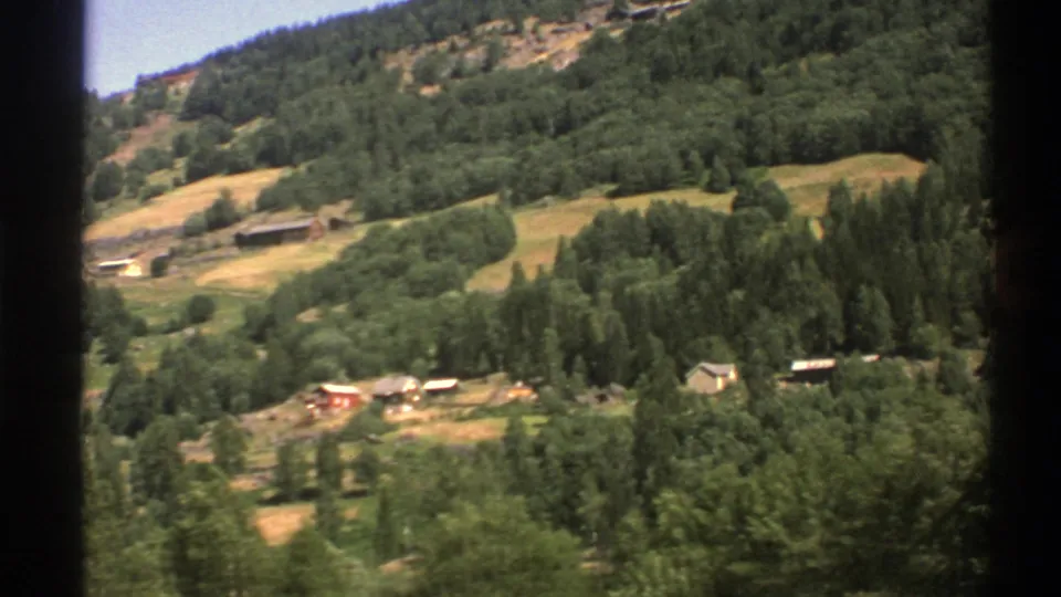 1976 Norway home-movie frame showing rural buildings and open fields across a green mountain slope.
