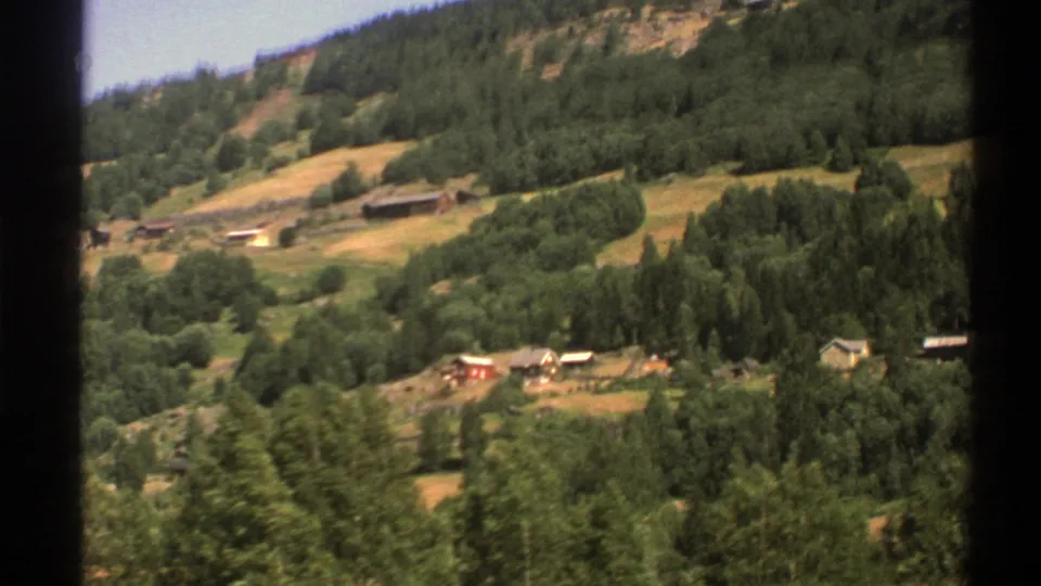 1976 8mm frame of a Norwegian hillside settlement with a red cabin, pale house, fields, and forest cover.