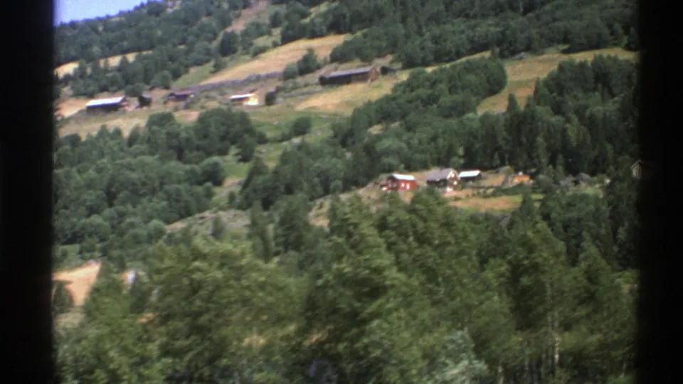 1976 Norway travel footage frame with a wooded valley foreground, hillside fields, and distant rural buildings.