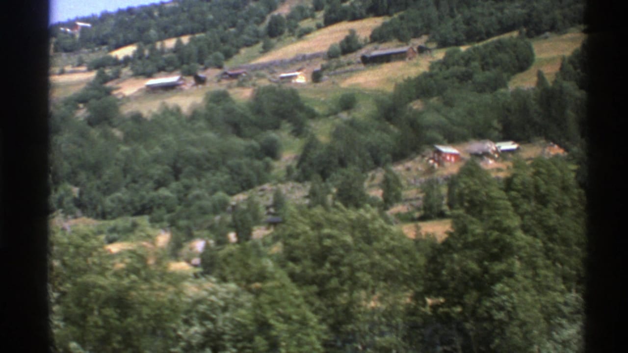 Distant rural hillside with fields, trees, and scattered buildings seen from a moving vehicle.