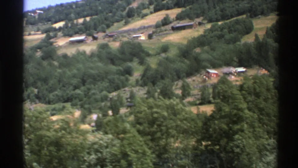 1976 8mm home-movie view of a Norwegian rural hillside with fields, trees, and small buildings seen through a moving-car frame.