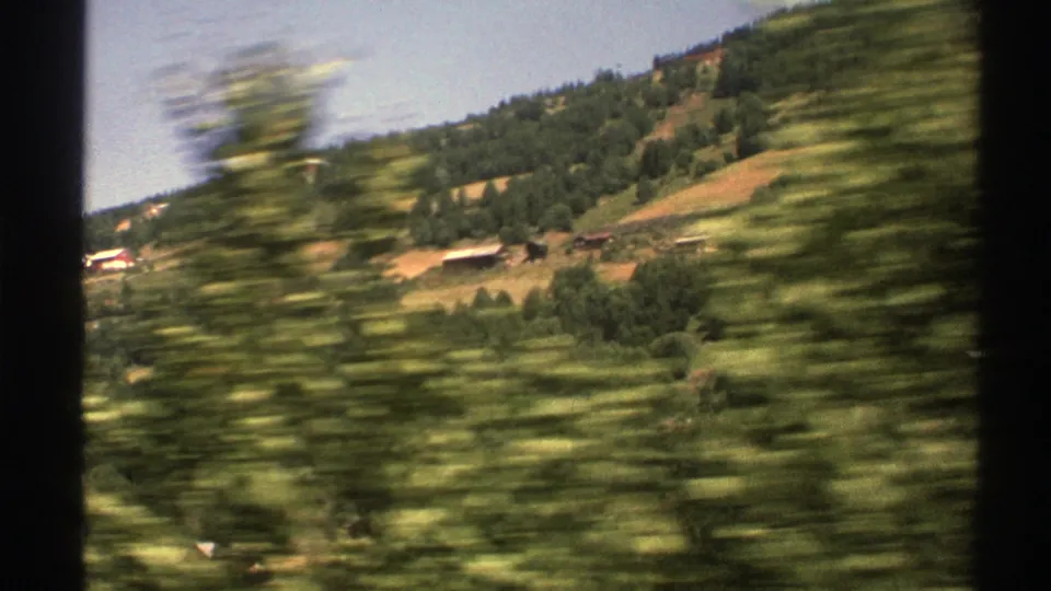 A 1976 Norwegian countryside view shows hillside fields and rural buildings glimpsed behind fast-moving foreground trees.