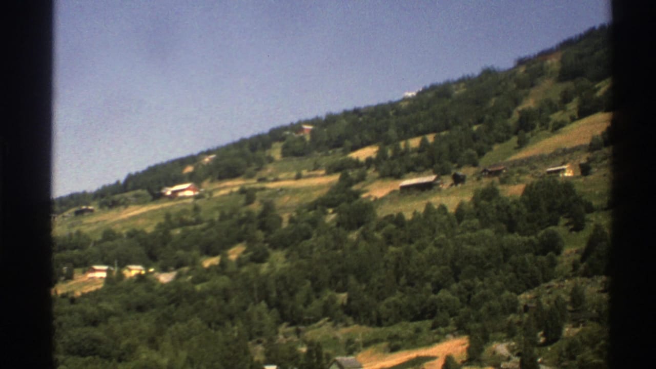 Green Norwegian hillside with scattered houses under a blue sky.