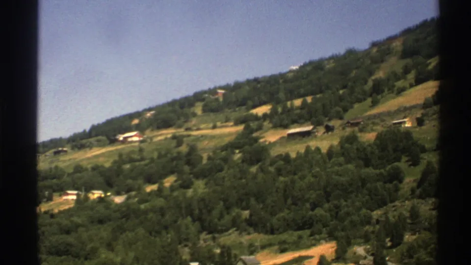 1976 8mm view of green hills in Norway, with scattered rural houses, forested slopes, and clear blue sky.