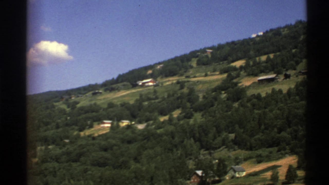 Norway 1976 home-movie frame showing a forested hillside, scattered houses, fields, and a small cloud in blue sky.