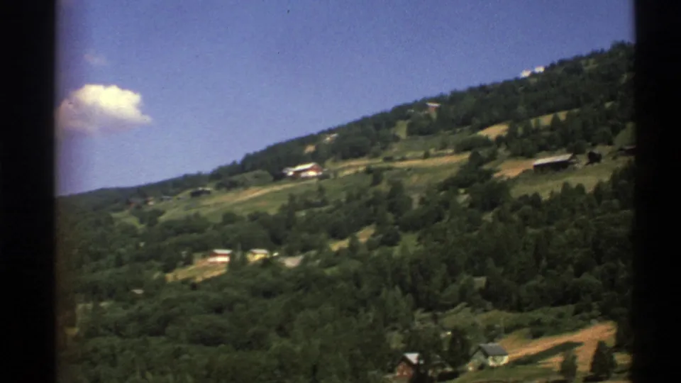 Norway 1976 home-movie frame showing a forested hillside, scattered houses, fields, and a small cloud in blue sky.