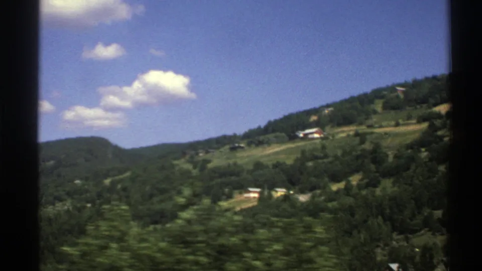 1976 Norway 8mm frame of a green valley and hillside with rural houses, fields, small clouds, and foreground motion blur.