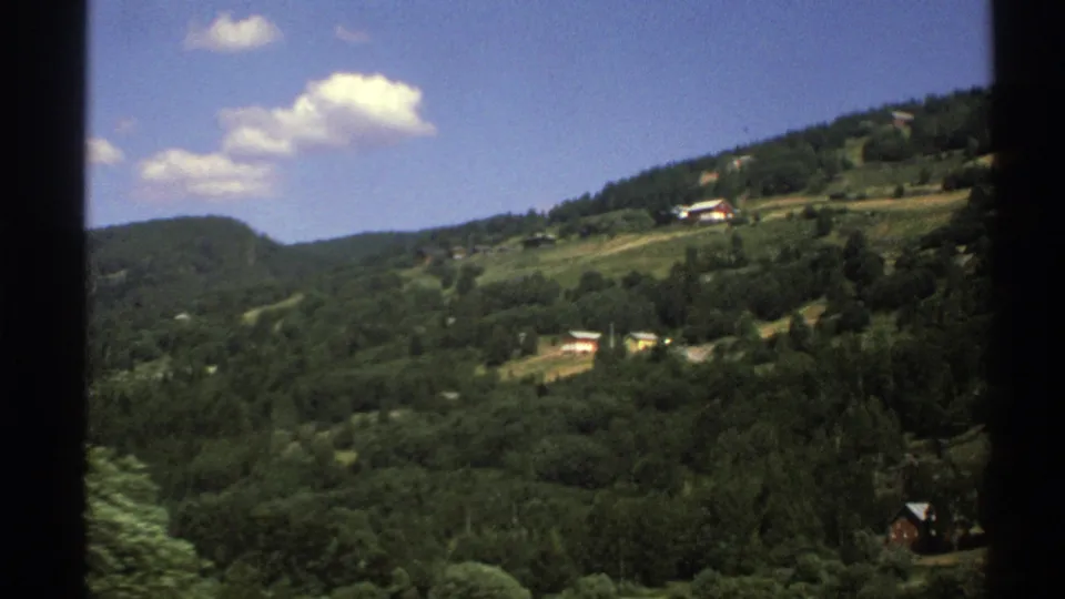 Norway 1976 home-movie view across wooded hills, scattered rural houses, pale fields, and small clouds in a clear sky.